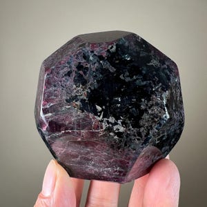 Pode incluir: Um cristal facetado, roxo escuro e preto, com uma superfície polida. O cristal tem uma forma geométrica complexa e exibe uma mistura de cores, incluindo roxos profundos e pretos. A pedra é segurada em uma mão.
