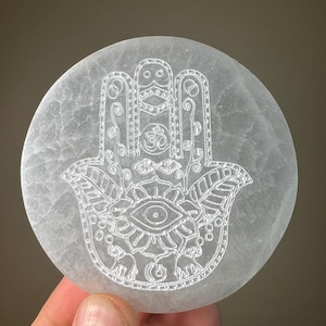 May include: A round, white selenite stone with a detailed etched Hamsa hand design. The design includes an eye, floral patterns, and the Om symbol. The stone is held in a hand.