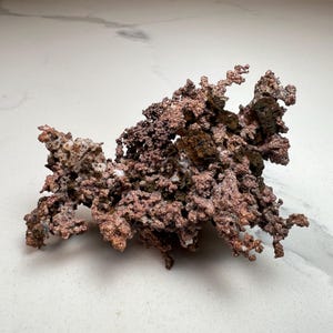 Pode incluir: Um esp&eacute;cime de cobre natural com uma estrutura ramificada complexa. A pe&ccedil;a exibe uma gama de cores, do castanho-avermelhado brilhante a tons mais escuros, quase pretos. A superf&iacute;cie tem uma apar&ecirc;ncia &aacute;spera e texturizada, com pequenas forma&ccedil;&otilde;es cristalinas.