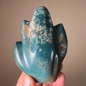May include: A teal and cream-colored stone sculpture with a rounded top and curved edges. The stone has a mottled appearance with areas of teal, cream, and brown. The sculpture is held in a hand.