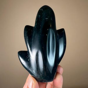 May include: A dark green, polished stone sculpture in the shape of a stylized cactus. The sculpture has a smooth, glossy surface with subtle variations in color and texture. The cactus-shaped sculpture is held in a hand.