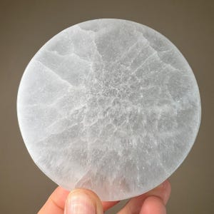 May include: A round, light gray stone coaster with a textured surface. The coaster is held by a hand and has a cloudy, marbled appearance. The stone is a pale, translucent color.