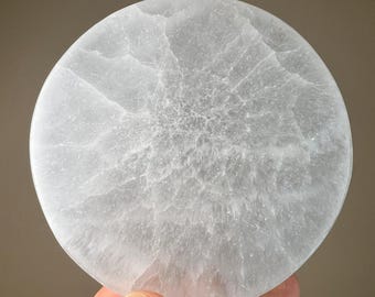 May include: A round, light gray stone coaster with a textured surface. The coaster is held by a hand and has a cloudy, marbled appearance. The stone is a pale, translucent color.