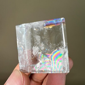May include: A clear, cube-shaped crystal with internal fractures. The crystal displays a rainbow iridescence on some surfaces, reflecting light in a spectrum of colors. The edges are sharp and well-defined, showcasing the crystal's geometric form.