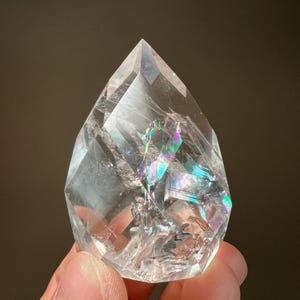 May include: A clear quartz crystal with a faceted, teardrop shape. The crystal has internal rainbows and light refractions. The crystal is held in a person's hand.