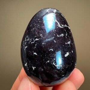 May include: A polished, egg-shaped gemstone with a deep purple hue and iridescent blue reflections. The stone has white veining throughout, adding texture to the smooth surface. The gemstone is held in a hand.