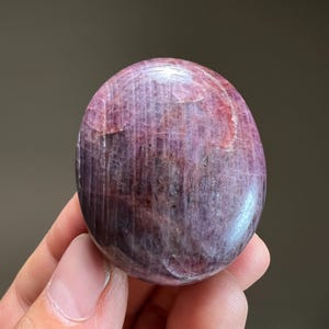May include: A polished, oval-shaped gemstone with a smooth surface, displaying a deep color palette of magenta, dark purple, and maroon. The stone features visible internal striations, natural mineral inclusions, and a glossy, reflective finish.