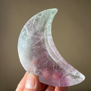 May include: A crescent moon shaped crystal carved from green and purple fluorite. The crystal is translucent and has a smooth, polished surface.
