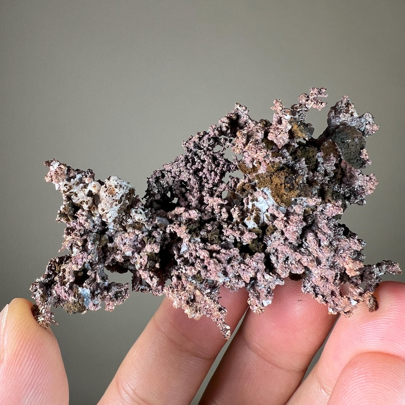 Pode incluir: Um esp&eacute;cime de cobre natural com uma estrutura ramificada complexa. O mineral exibe uma mistura de tons avermelhados-castanhos, viol&aacute;ceos e acinzentados, com uma superf&iacute;cie &aacute;spera e texturizada. O esp&eacute;cime &eacute; segurado entre os dedos.