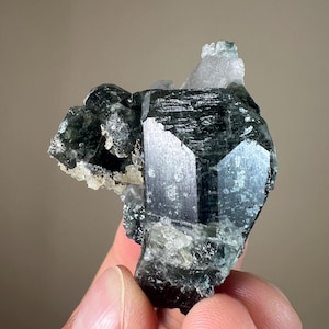May include: A cluster of dark green and black crystals with a glassy sheen. The crystals are arranged in a cluster, with some of the crystals being more prominent than others. The crystals are set against a light background.