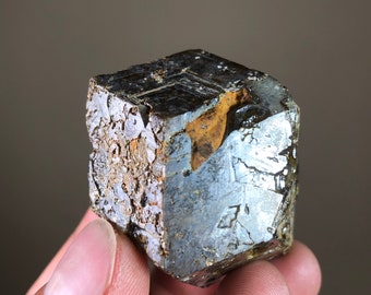 May include: A dark gray, metallic, cube-shaped crystal with a slightly iridescent sheen. The crystal has a rough, textured surface and appears to be a natural mineral specimen.