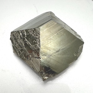 May include: A large, faceted pyrite crystal with a metallic, golden-silver sheen. The mineral specimen has a geometric shape with multiple flat surfaces and visible striations. The rough, natural edges contrast with the polished facets, highlighting its crystalline structure.
