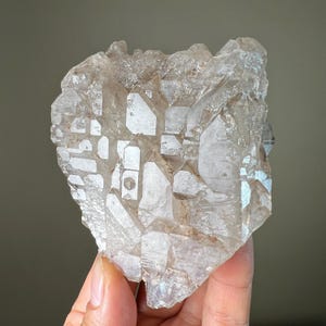 May include: A large, clear quartz crystal cluster with multiple points and facets. The crystal has a translucent appearance with subtle variations in color, ranging from clear to light brown. The surface reflects light, creating a shimmering effect. The overall shape is irregular and natural.