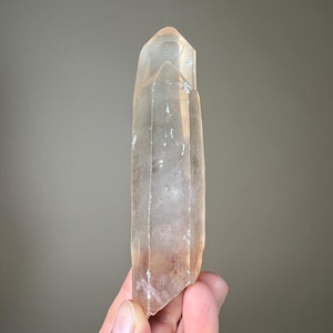 May include: A clear quartz crystal with a light orange hue. The crystal is held in a hand and has a pointed tip. The surface has some imperfections. The crystal is a natural mineral specimen.