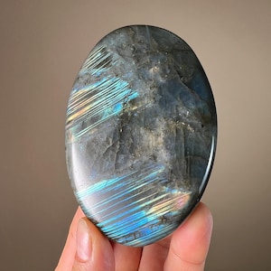 May include: Oval-shaped labradorite gemstone with a polished surface. The stone displays iridescent flashes of blue, green, and yellow across a gray and black base. The gemstone is held in a hand, showcasing its natural beauty and unique color variations.