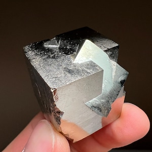 May include: A cubic pyrite crystal with a metallic, silver-like luster. The crystal has a complex geometric shape with multiple facets and a reflective surface. The mineral is held in a hand against a dark background.