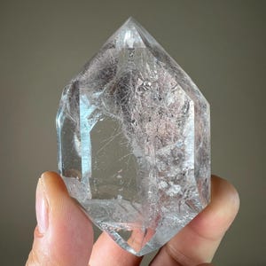 May include: A clear quartz crystal with a pointed top and multifaceted sides. The crystal has internal inclusions and is held in a hand. The quartz is transparent, allowing light to pass through, and is a natural mineral specimen.