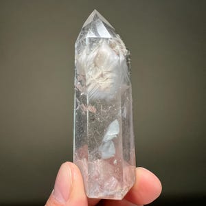 May include: A clear quartz crystal point with internal inclusions. The crystal is transparent with a pointed top and a rectangular base. The inclusions appear as white and grey wisps within the crystal. The crystal is held by a person's fingers.