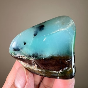May include: A polished gemstone with a striking color gradient. The top portion is a vibrant turquoise, transitioning to a deep brown base. Black inclusions add detail. The stone is held in a hand, showcasing its smooth, glossy surface.