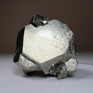 May include: A pyrite crystal cluster with a multifaceted, geometric structure. The surface has a reflective, silvery sheen with visible cracks and imperfections. The crystal's shape is irregular, with multiple facets and a dark, contrasting top.