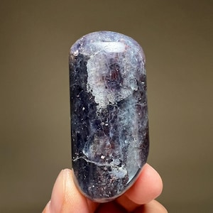 May include: A polished, elongated stone with a rounded top. The stone features a blend of deep purple, white, and hints of red, creating a marbled effect. The surface is smooth and reflective, with natural variations in color and texture.