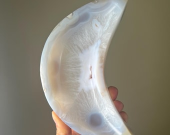 May include: A crescent-shaped agate stone with a polished surface. The stone features a mix of white, cream, and translucent areas, with a central, lighter-colored pattern. The overall shape is smooth and curved, with a natural, earthy aesthetic.