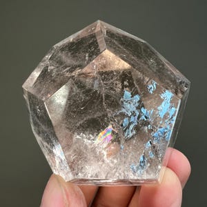 May include: A clear, faceted quartz crystal with a pentagonal shape. The stone features internal fractures and inclusions that create reflective surfaces, including a small, distinct rainbow-colored prism effect visible within the transparent structure.