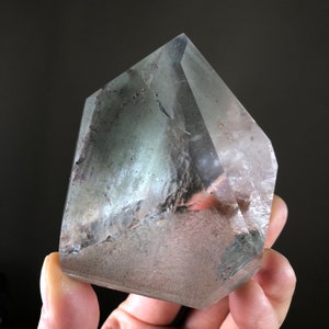 May include: A clear, faceted crystal with a green and white cloudy interior. The crystal is shaped like a pyramid with a pointed top and a flat base.