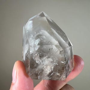 May include: A clear quartz crystal point with a faceted, geometric form. The crystal has internal inclusions and a smoky, translucent appearance. The point is held by a person's fingers.