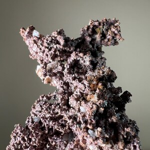 Pode incluir: Close-up de um esp&eacute;cime mineral de cobre com uma superf&iacute;cie &aacute;spera e texturizada. O mineral tem uma mistura de cores castanho-avermelhadas e castanho-escuras, com pequenas inclus&otilde;es de cristais brancos. A forma geral &eacute; irregular e irregular.