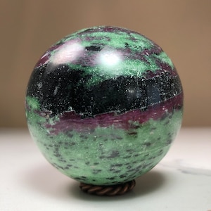 May include: A green, black, and purple gemstone sphere on a copper stand. The sphere is polished and has a smooth surface.