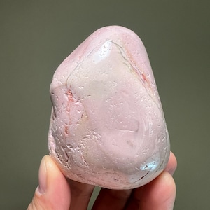 May include: A smooth, rounded pink stone with subtle variations in color and texture. The stone has a polished appearance, with areas of lighter and darker pink hues, and some reddish veins. The overall shape is irregular, with a rounded top and a slightly flattened base.