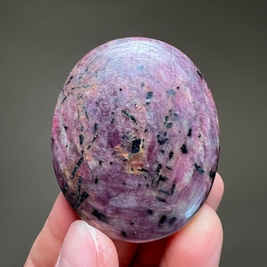 May include: Oval-shaped polished gemstone with a mix of pink and purple hues, speckled with black inclusions. The stone has a smooth, rounded surface and a natural, organic appearance. The gemstone is held in a hand.