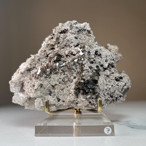 May include: A rough, gray mineral specimen with dark, reflective crystal formations. The mineral is mounted on a clear acrylic base with a gold-colored metal support. The base has a small white sticker with the number 7.