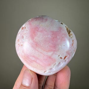 May include: A polished, oval pink gemstone with white and darker pink veining. The stone has a smooth, glossy surface and a few small natural holes. The gemstone is held in a hand.