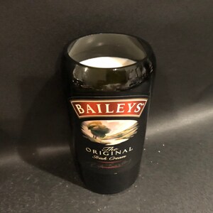 May include: A black glass with the label of Baileys The Original Irish Cream. The glass is filled with a white liquid. The label features the Baileys logo and a landscape scene.