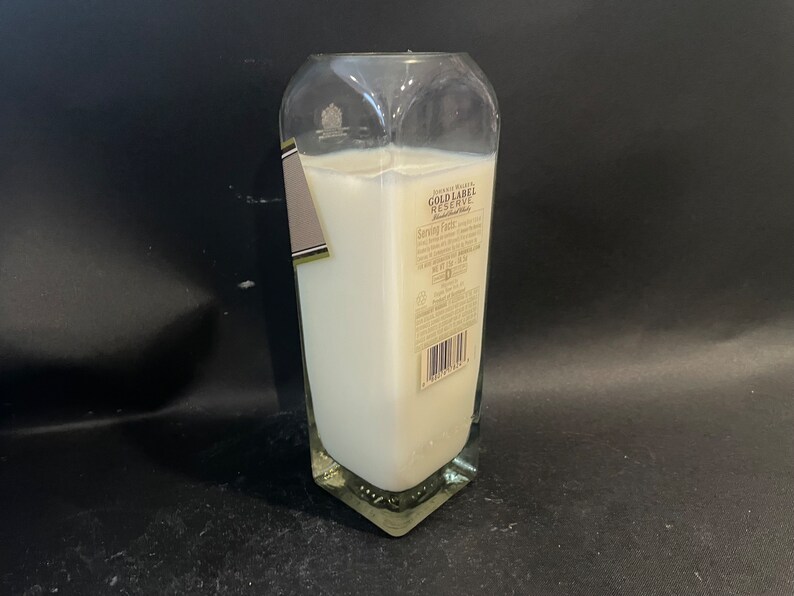 May include: A clear glass candle jar with a white candle inside. The jar is shaped like a liquor bottle and has a label that reads "Johnnie Walker Gold Label Reserve Blended Scotch Whisky".