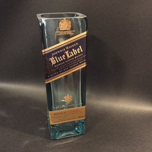 May include: A rectangular, clear glass bottle with a blue tint. The bottle features a label with the text "Johnnie Walker Blue Label Blended Scotch Whisky". The label is primarily navy blue and gold. The bottle is empty.
