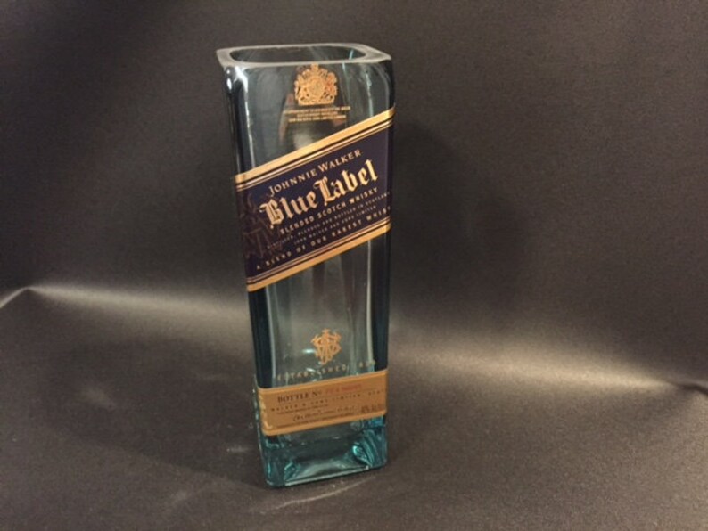May include: A rectangular, clear glass bottle with a blue tint. The bottle features a label with the text "Johnnie Walker Blue Label Blended Scotch Whisky". The label is primarily navy blue and gold. The bottle is empty.