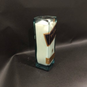 May include: A rectangular glass candle holder with a light blue tint, filled with white wax. The candle holder has a label that reads "Blue Label" in gold and navy blue. The candle holder is approximately 20 cm tall.