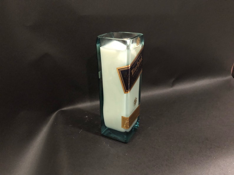 May include: A rectangular glass candle holder with a light blue tint, filled with white wax. The candle holder has a label that reads "Blue Label" in gold and navy blue. The candle holder is approximately 20 cm tall.