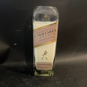 May include: A clear glass candle in the shape of a Johnnie Walker Gold Label Reserve whisky bottle. The label is gold with black text and a walking man logo.