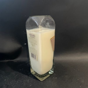 May include: A clear glass bottle candle with a white label that reads "Gold Label Reserve" and a barcode. The candle is lit and the wax is melted.