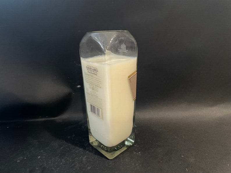 May include: A clear glass bottle candle with a white label that reads "Gold Label Reserve" and a barcode. The candle is lit and the wax is melted.