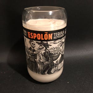 May include: A clear glass candle jar with a white wax candle inside. The jar has a black and white label with the text "Espolón Tequila Reposado" and a graphic of skeletons carrying a basket of fruit.
