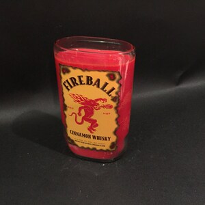 May include: A red glass candle with a Fireball Cinnamon Whisky label. The label features a red dragon with the words "Fireball" and "Hot" on it.