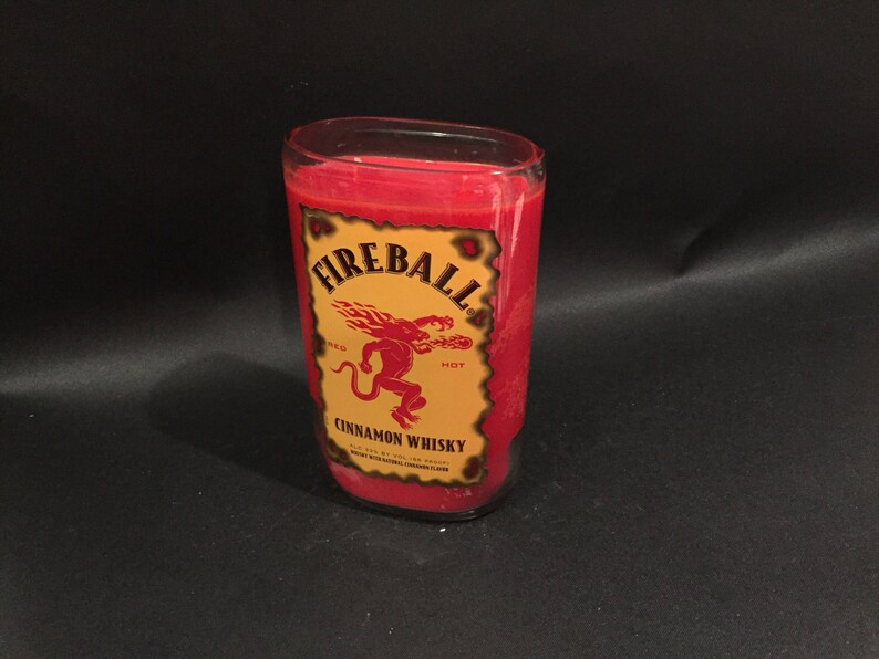 May include: A red glass candle with a Fireball Cinnamon Whisky label. The label features a red dragon with the words "Fireball" and "Hot" on it.