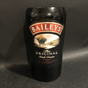 May include: A black glass with the Baileys logo and text. The logo is in a red and white frame, with an image of a landscape. The text reads "The Original Irish Cream".