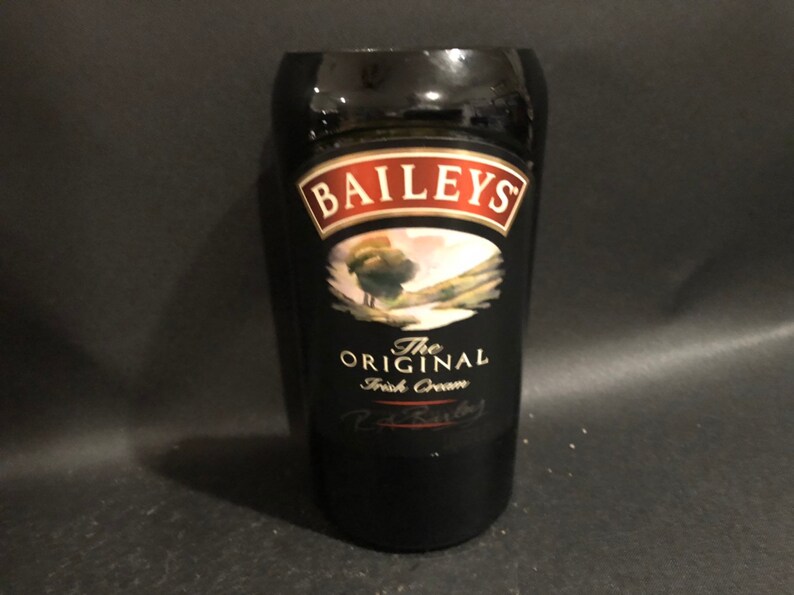 May include: A black glass with the Baileys logo and text. The logo is in a red and white frame, with an image of a landscape. The text reads "The Original Irish Cream".