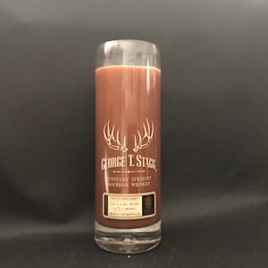 May include: A clear glass candle with a brown wax scent. The candle has a label with the text "George T. Stagg Kentucky Straight Bourbon Whiskey" and a stag head logo.